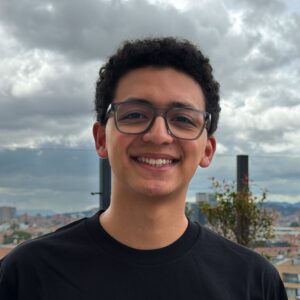 Smiling young man with curly hair and gray-framed glasses, wearing a black shirt, outside on a rooftop with a cloudy sky and city skyline in the background.