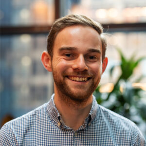 Portrait of a smiling man with short blond hair and a beard, wearing a blue-and-white checkered shirt in a bright indoor setting, possibly an office.