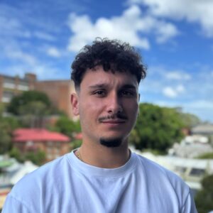 Portrait of a young man with short curly hair and light stubble wearing a white T-shirt, outdoors on a sunny day with blue skies behind him.