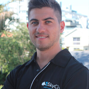 Smiling man in a black SkyCity polo shirt, outdoors with buildings and greenery in the background.