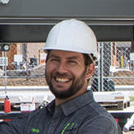 Trabajador de la construcción con un casco blanco sonríe a la cámara en un lugar de trabajo con cercas y equipos en el fondo.
